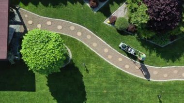 Güzel ve manikürlü yeşil bahçeyle ilgilenen adam. Eğri kaldırım boyunca yuvarlak basamaklı çim biçen işçinin hava görüntüsü. 