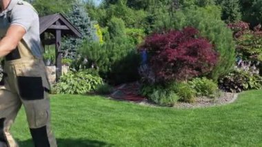 Çim biçme makinesiyle geniş yeşil çimenleri budamaya çalışan erkek işçiyi yakından görebiliriz. Konut Bahçesi ve Peyzaj Mevsimsel Bakım.