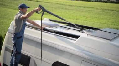 Erkek karavan sahibi, arabanın çatısını basınç çubuğuyla temizliyor. Güneş panellerine ve tavan penceresine dikkatlice su püskürtüyor.. 