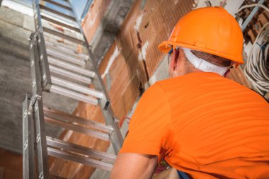 Kafkas İnşaat İşçisi 40 'lı yaşlarda Alüminyum Merdiven kullanarak yukarı çıkan Turuncu Sert Şapkalı. İnşaat Sitesi TemasıName.