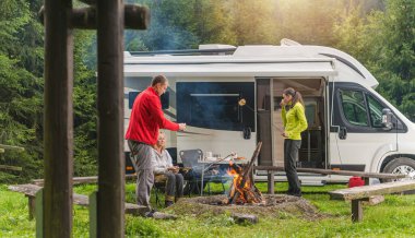 Three Caucasian Friends in Their Late 30s, One Men and Two Woman Having Fun on Wilderness Camping with Modern RV Camper Van. Grilling Bread and Polish Sausage on Campfire. clipart