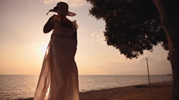 60代の白いドレスとサンハットを着た引退した女性が夏の海の夕日を楽しむ — ストック動画