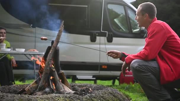 40代の白人笑顔の男性キャンプファイヤーでのポーランド料理のソーセージと彼の配偶者との会話を作る キャンパー ヴァンとの休暇時間 — ストック動画