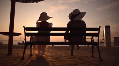 Büyük Güneş Şapkalı İki Zarif Beyaz Kadın Denizin ve Günbatımının tadını çıkarıyor