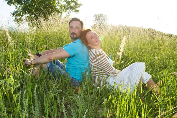 Couple in big green grass back to back. Man and woman sitting in the field in summer day. Rustic loving couple gently peacefully gossiping sits back to back in big field grass