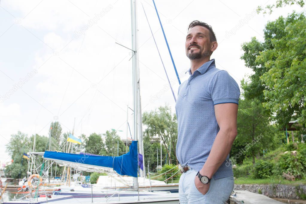 Businessman spending free time walking on marina and looking sightseeing during summer vacation. Portrait of young happy yachtsman in marina near of boats and green bank