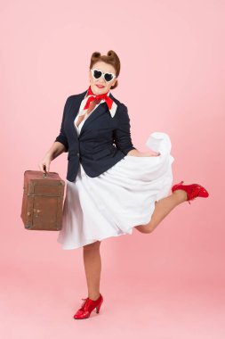 Bavul ve siyah güneş gözlüğü ile komik gezgin kız. Pin-up tarzı vintage dava ile esmer kadın. Genç kırmızı topuklu pembe arka plan izole mutlu yolculuk Bayan gülümsedi. Pin-up Bayan atlama.