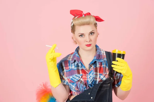 Girl in pin-up style drink coffee and holds cigarette in hands. Coffee break after cleaning. Housewife in yellow rubber cloves with soft duster and coffee in hand. Repair girl on pink background