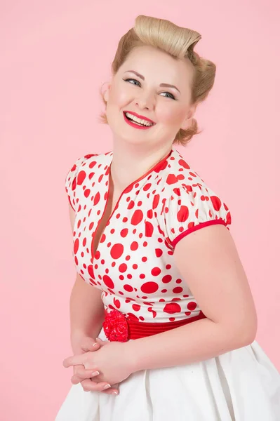 Pin-up vintage makyaj duygu güven elele ve komik kameraya bakarak mutlu gülümsüyor çekici kadın. Stüdyo pastel pembe arka plan üzerinde beyaz kırmızı nokta elbiseli sarışın curl kadın