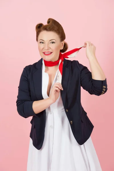 Brunette woman with red scarf on neck. Air uniform for girls on airplane. Happy girl with pin-up style and make-up in blue jacket take on the red scarf on pink background.