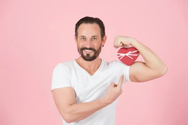 Man with red heart. Smiling guy in white t-shirt and present box. Bearded guy holds heart-box on bicep on rose background. Happy man pointing on box with present. Valentine day surprise.