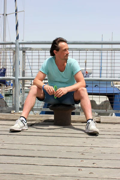 man in t-shirt sitting on bollard on pier with barrier in marina with boats