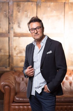 Gözlük takan ve ceketini arka planda kahverengi kanepe ile dururken dokunmadan düşünceli orta yaşlı adam