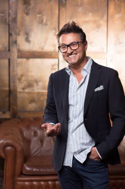 Kahverengi kanepe önünde duran ve süre hareketleri kullanarak ve mesafe içine bakarak gülen neşeli duygusal zarif adam