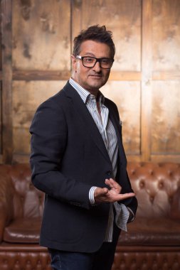 Etkileyici Avrupa adam arka plan üzerinde kahverengi kanepe ile ayakta ve onun parmak işaret ederken gülümseyen şık elbiseli