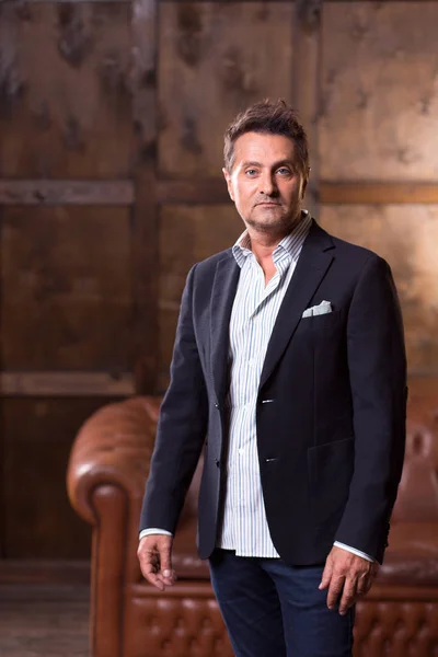 Thoughtful middle aged man in dark suit standing alone with brown sofa on the background and looking at you