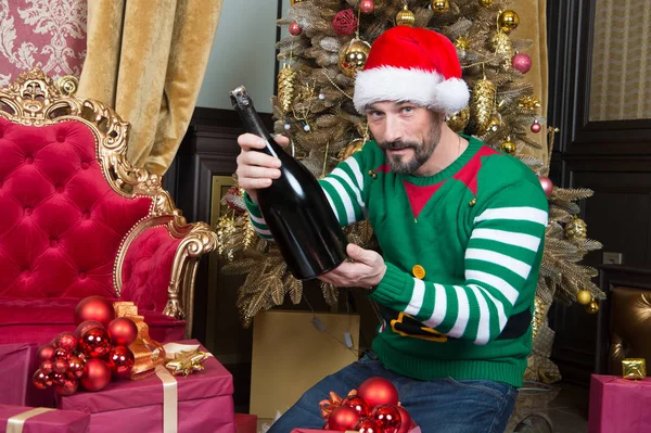 Relaxed funny man in lovely elf costume sitting near the Christmas tree surrounded by presents and holding a big bottle