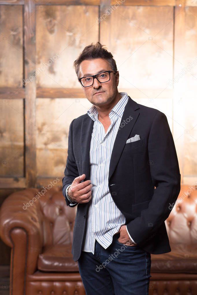Thoughtful middle aged man wearing glasses and touching his jacket while standing with the brown sofa on the background