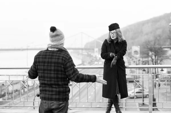 Man in flannel jacket delayed to date. Woman shows to watches on arm. Man spread his arms from the strong woman. Delayed dated on bridge. Man and woman on bridge. Strong lady in glasses confirm late time for man in hat.