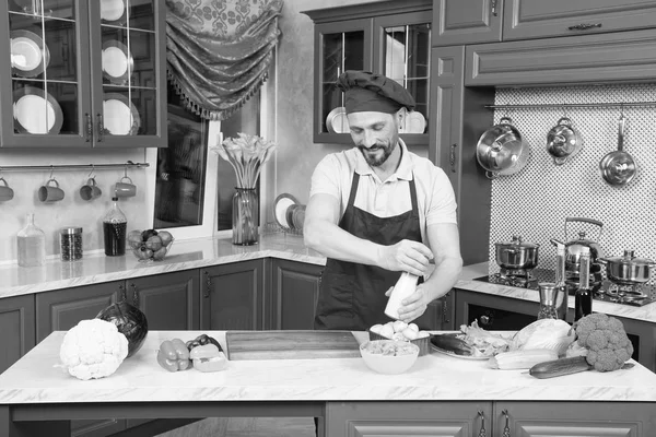Smiling bearded cook in apron and chef cap standing at kitchen table while salting vegetable salad and expressing kindness on his face