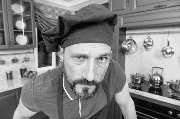 Close up of confused bearded chef in kitchen uniform looking at you with suspicion while keeping his right eye half open