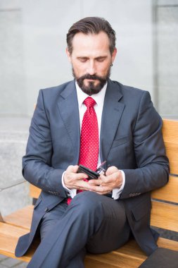 Gri takım elbise giyiyor ve cep telefonuyla ve onun arkadaşlarından iletileri okuma sırasında tahta bankta oturan yakışıklı işadamı sakin sakallı