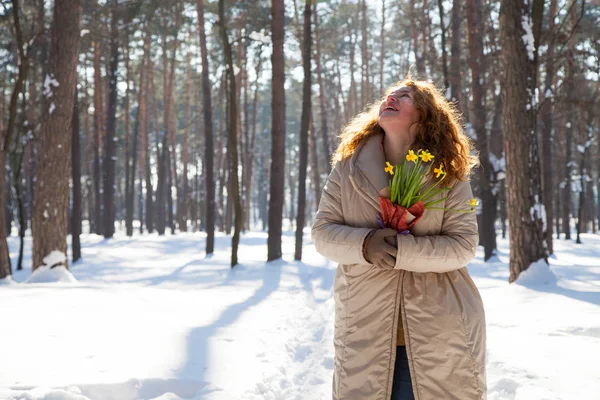 High spirits. Optimistic woman with red curly hair keeping yellow narcissus in hands while looking up during pleasant walk through winter forest