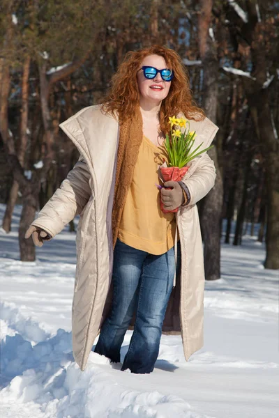 Fashionable smiling woman in unzipped coat keeping yellow narcissuses in hand while walking through snow in winter forest