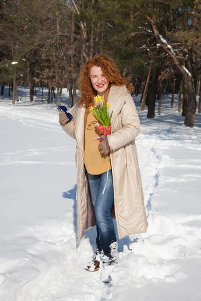 Full length of pleased charming woman in winter coat holding yellow narcissuses while walking along snowy footpath and looking at you with kind hearted smile