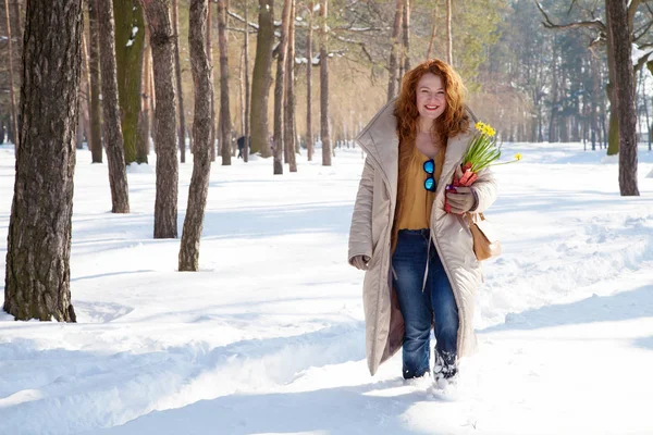 Full of positive emotions. Optimistic red haired woman expressing excitement while holding yellow narcissuses and going through snowdrifts in sparse forest