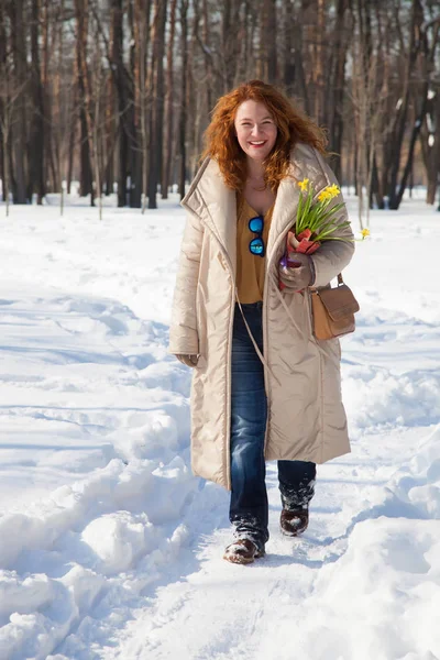 Full length of positive smiling woman in warm clothes holding yellow narcissuses and walking along footpath against sparse winter forest
