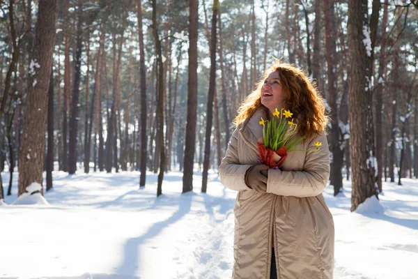 Joyful middle aged woman expressing curiosity while looking up and holding bouquet of yellow narcissus in hands against winter forest landscape