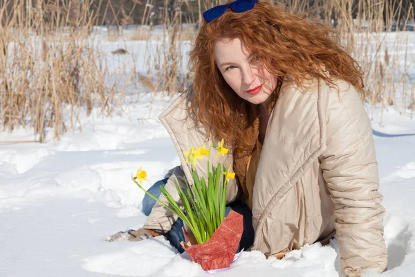 Charming glance. Gorgeous red haired woman sitting on snow surface and looking at you while expressing calmness with yellow narcissus on foreground