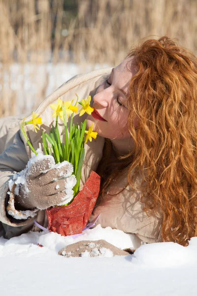 Vertical image of restful smiling woman lying in snow while smelling yellow narcissus and being pleased