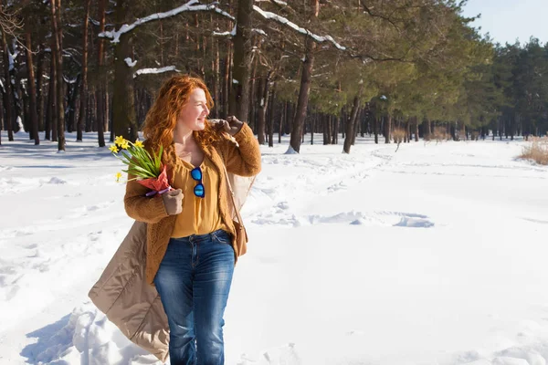 Joyful red haired woman looking into the distance with her coat over shoulder and bouquet of yellow narcissus in hand while walking through snowdrifts with pine trees on the background