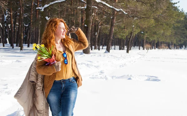 Relaxation. Horizontal image of pleasant red haired woman having a stroll through forest snowdrifts while holding her coat over shoulder and restfully looking into the distance