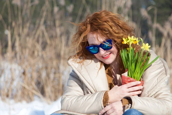 Pleased adorable woman having rest in snow while keeping bouquet of yellow narcissus in hand and looking down through fashionable blue sunglasses with smile on her face