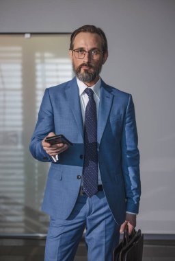 Ofis binasında akıllı telefon kullanan yaşlı bir profesyonel. İş adamı cep telefonu tutuyor. Uygulama mesajları kullanıyor. Ceket ve çanta takıyor..