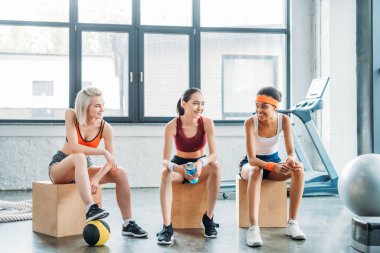 Birbirinizle konuşmak ve spor salonunda ahşap kutular üzerinde oturan mutlu çok ırklı kadın sporcular