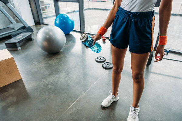 cropped shot of sportswoman in wristbands standing with bottle of water at gym