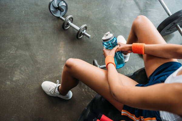cropped image of sportswoman in wristbands sitting with bottle of water on training tire at gym 
