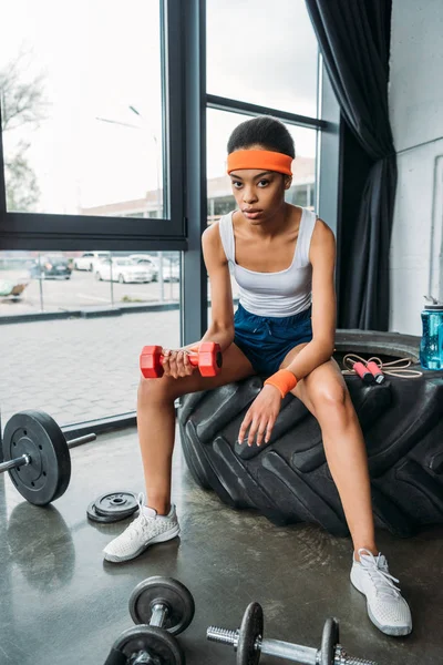 african american sportswoman in headband and wristbands doing exercise with dumbbell while sitting on training tire at gym
