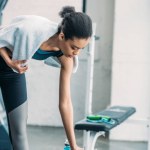 African american sportswoman with towel taking sportive water bottle after workout at gym