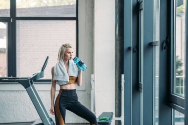 young sportswoman with towel drinking water from sportive water bottle after workout at gym clipart