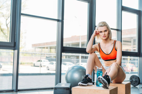 young attractive sportswoman with sportive water bottle resting on wooden boxes after work out at gym