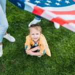Recortado disparo de padres sosteniendo bandera americana y poco hijo agachándose en la hierba