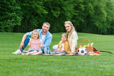 birlikte piknik parkta otururken kameraya gülümseyen iki çocuklu mutlu aile 