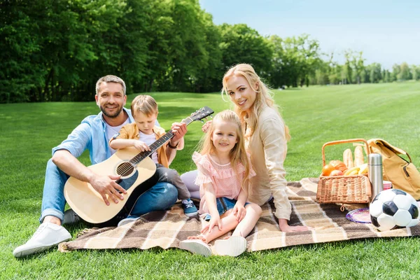 愉快的家庭与吉他微笑在照相机 而坐在一起在格子在野餐 — 图库照片