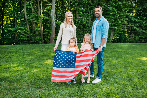 счастливая семья с двумя детьми, держащая в руках наш флаг и улыбающаяся фотоаппарату в парке
  