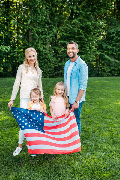 happy family with two kids holding american flag and smiling at camera in park  